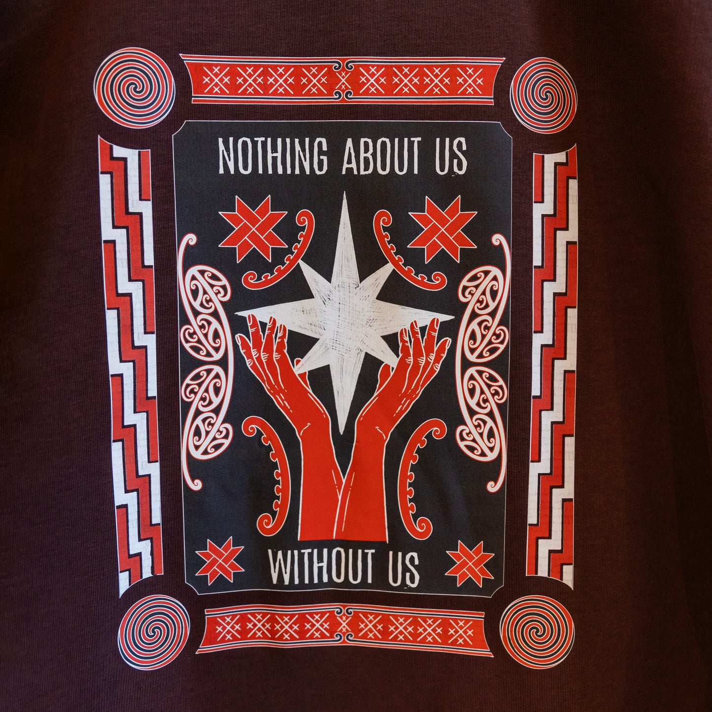 A close up of the print on a brown sweatshirt with a bold design reading "Nothing About Us Without Us." The graphic features red hands, stars, koru and spirals.