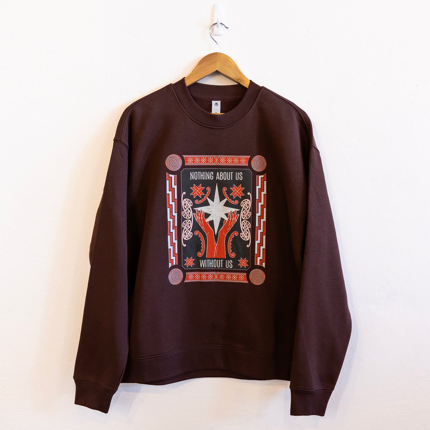 A brown sweatshirt with a bold design reading "Nothing About Us Without Us" hung up on a coat hanger against a white wall. The graphic features red hands, stars, koru and spirals.