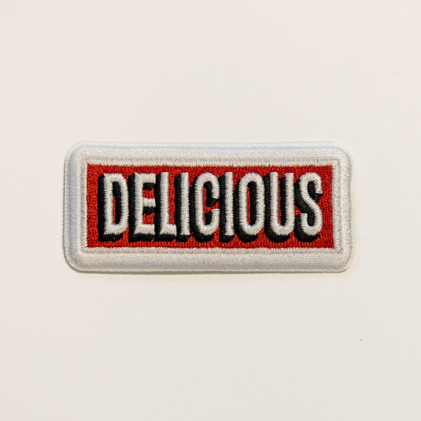 Rectangular embroidered patch with the word "DELICIOUS" in bold white and black letters on a red background, framed by a white border.