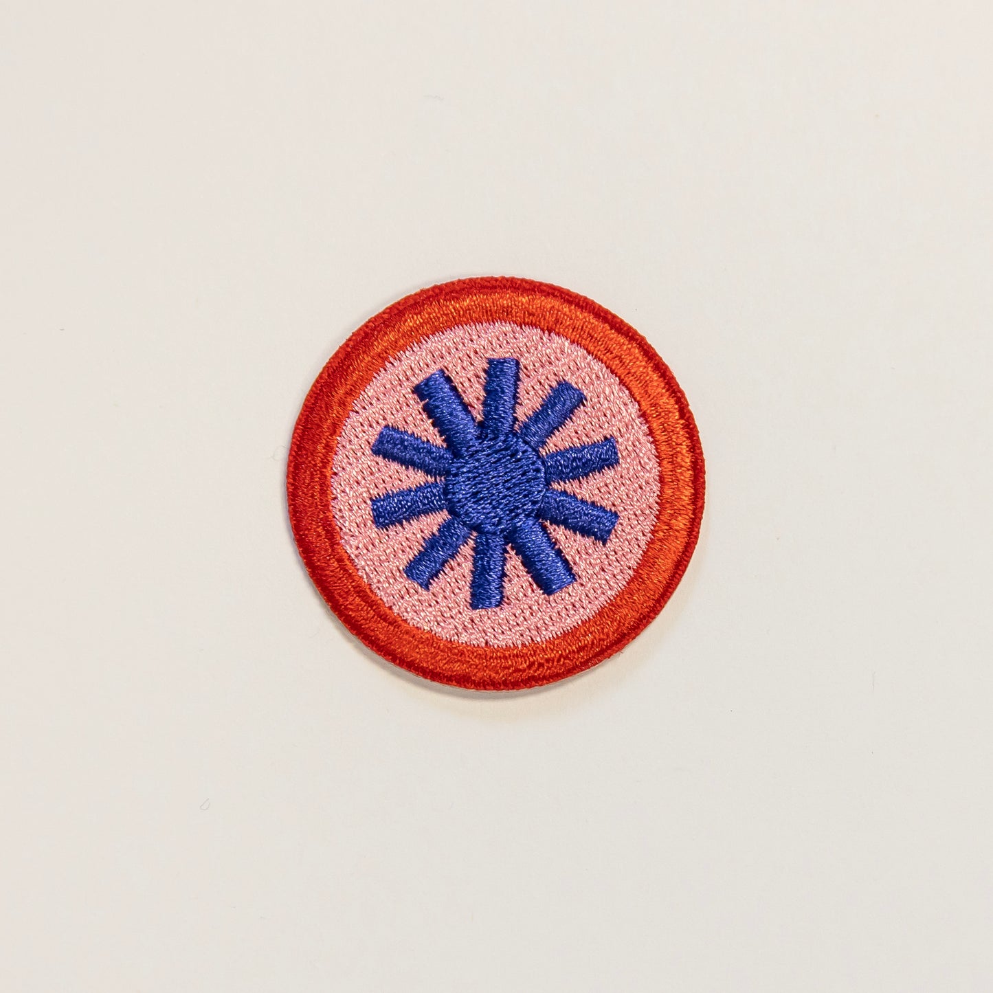 Circular embroidered patch on a light background, featuring a blue tohuwhetū symbol at the center, surrounded by a pink field and red border.