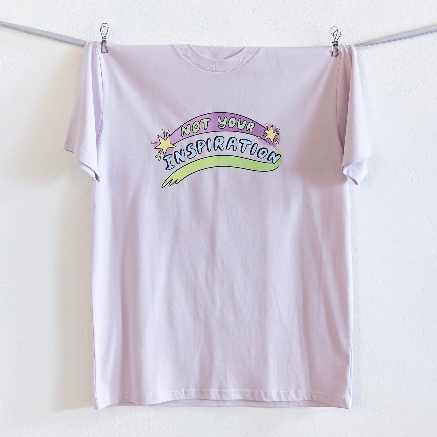 A light pink T-shirt with the phrase "Not Your Inspiration" in colourful letters is hung up on a silver ribbon against a white wall. The words 'Not your' appear in lime green on a purple swoosh, positioned between two yellow stars. Beneath these words, 'inspiration' appears in all caps, while a second stroke of colour, which appears like a green-yellow paint stroke, provides a boundary of support at the base of the print.