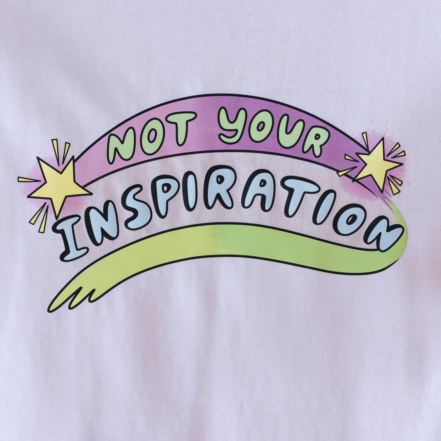 Close up photo of a light pink T-shirt with the phrase "Not Your Inspiration" in colourful letters. The words 'Not your' appear in lime green on a purple swoosh, positioned between two yellow stars. Beneath these words, 'inspiration' appears in all caps, while a second stroke of colour, which appears like a green-yellow paint stroke, provides a boundary of support at the base of the print.