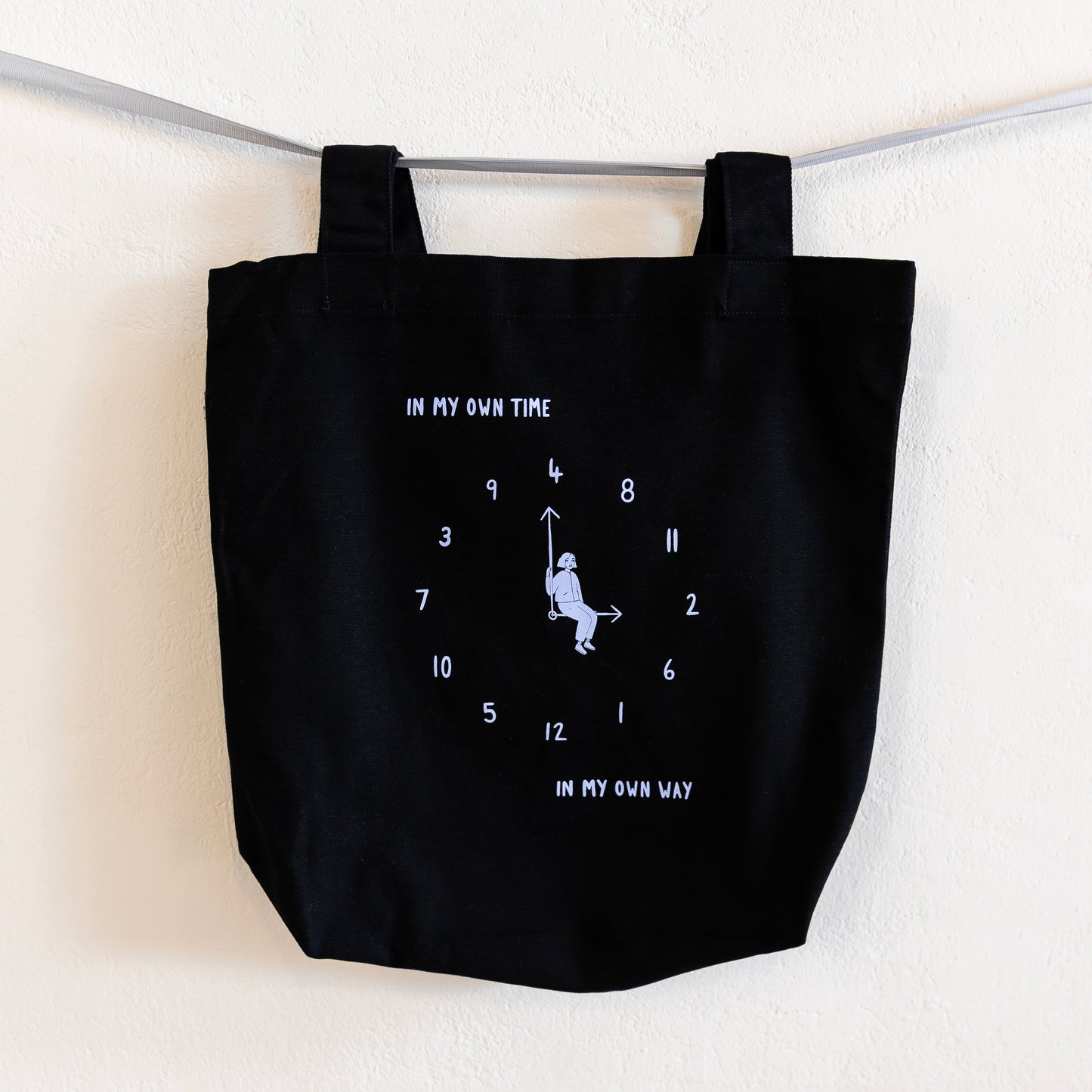 A black tote bag with a light blue print is hung on a silver ribbon against a white wall. The design features an analogue clock, with jumbled numbers and a person sitting on the clock hands. Above the design reads "IN MY OWN TIME" and below reads "IN MY OWN WAY".