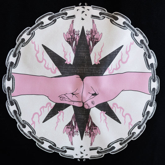 A close up of the design on the black tote bag. The central circular design is of two pink fists bumping inside a chain circle with breaks at the top and bottom. The inside of the circle has a beige background, and the fists are in-front of a black star with pink flames and flowers branching off of it.