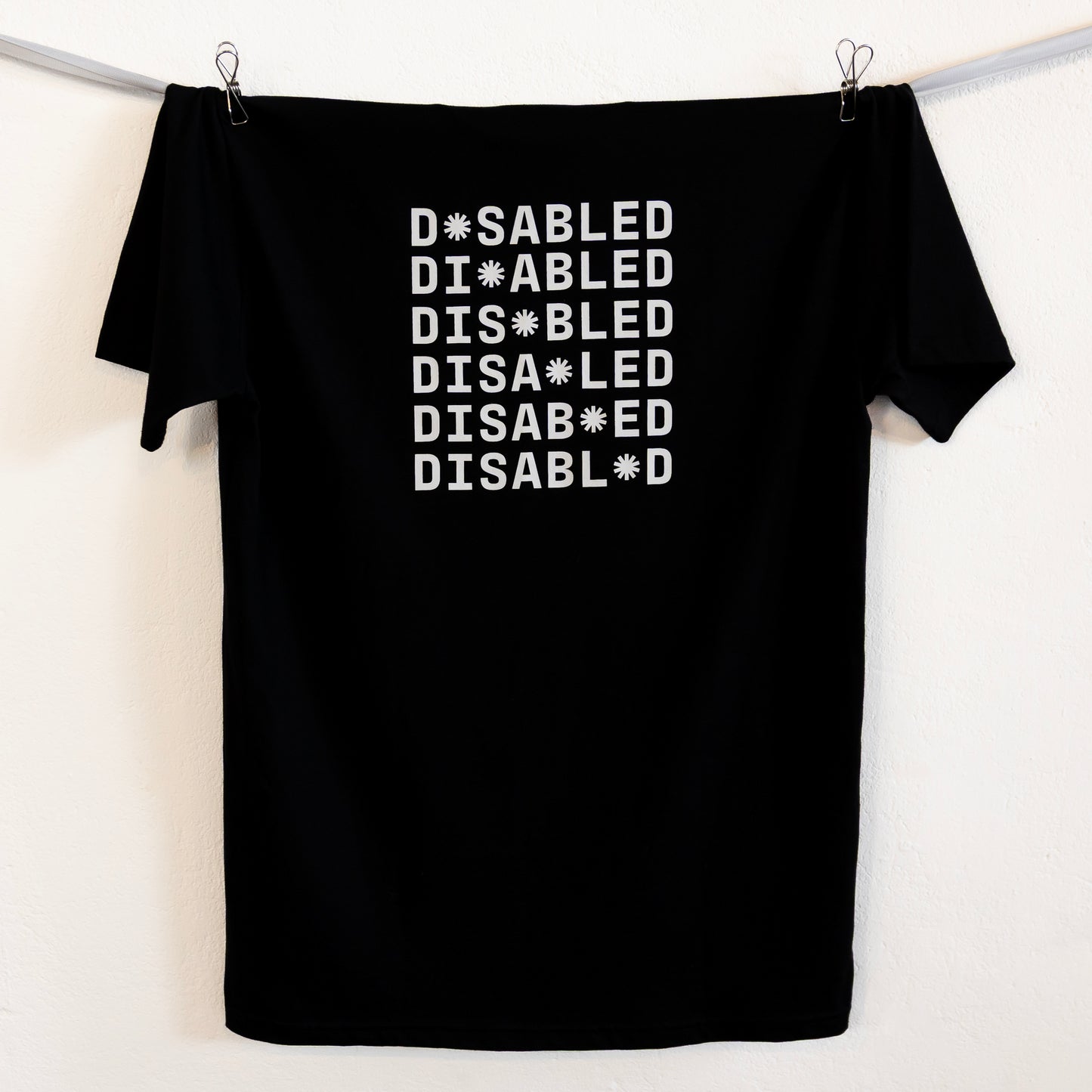 Black t-shirt hung up on a silver ribbon against a white background. The tee features the word "DISABLED" repeated six times in white with a tohuwhetū replacing different letters from left to right on each line.