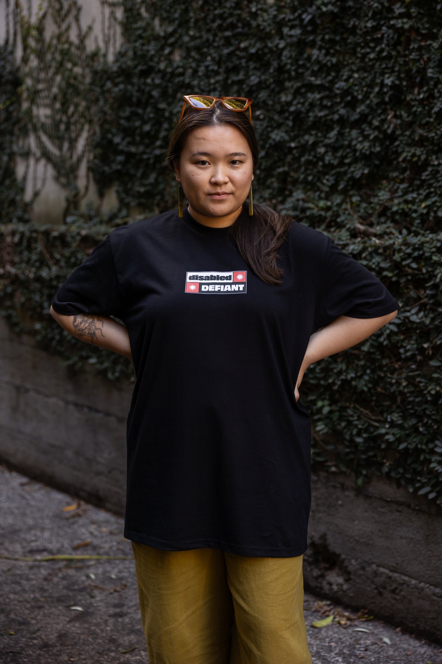 Eda stands in-front of a leafy wall outdoors, wearing a black T-shirt, untucked, with "disabled, DEFIANT" in bold text stands outdoors. The design is black, red and white, and features two tohuwhetū.