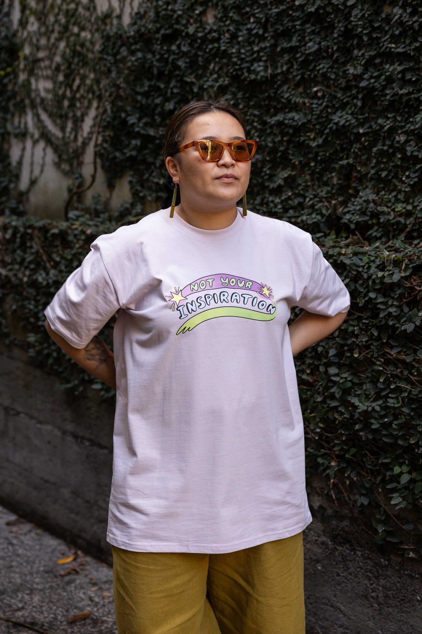 Eda wearing a light pink T-shirt with the phrase "Not Your Inspiration" in colourful letters stands against a leafy background. The words 'Not your' appear in lime green on a purple swoosh, positioned between two yellow stars. Beneath these words, 'inspiration' appears in all caps, while a second stroke of colour, which appears like a green-yellow paint stroke, provides a boundary of support at the base of the print.