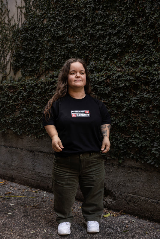 Ella stands in-front of a leafy wall outdoors, wearing a black T-shirt with "disabled, DEFIANT" in bold text stands outdoors. The design is black, red and white, and features two tohuwhetū.