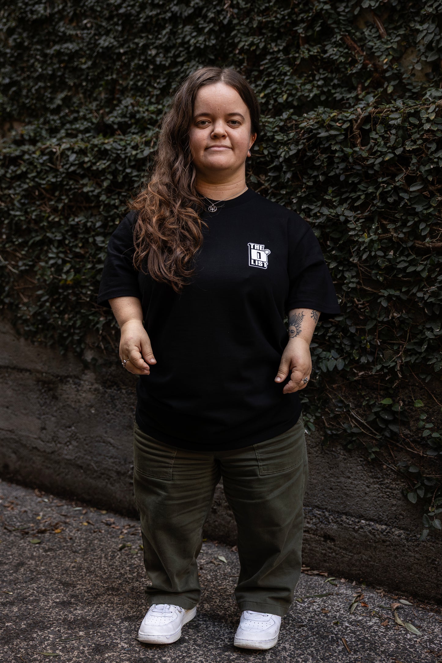 Ella stands outdoors against a wall covered with green ivy. She wears a black T-shirt with a white "The D*List" logo on the left chest, olive pants, and white shoes.