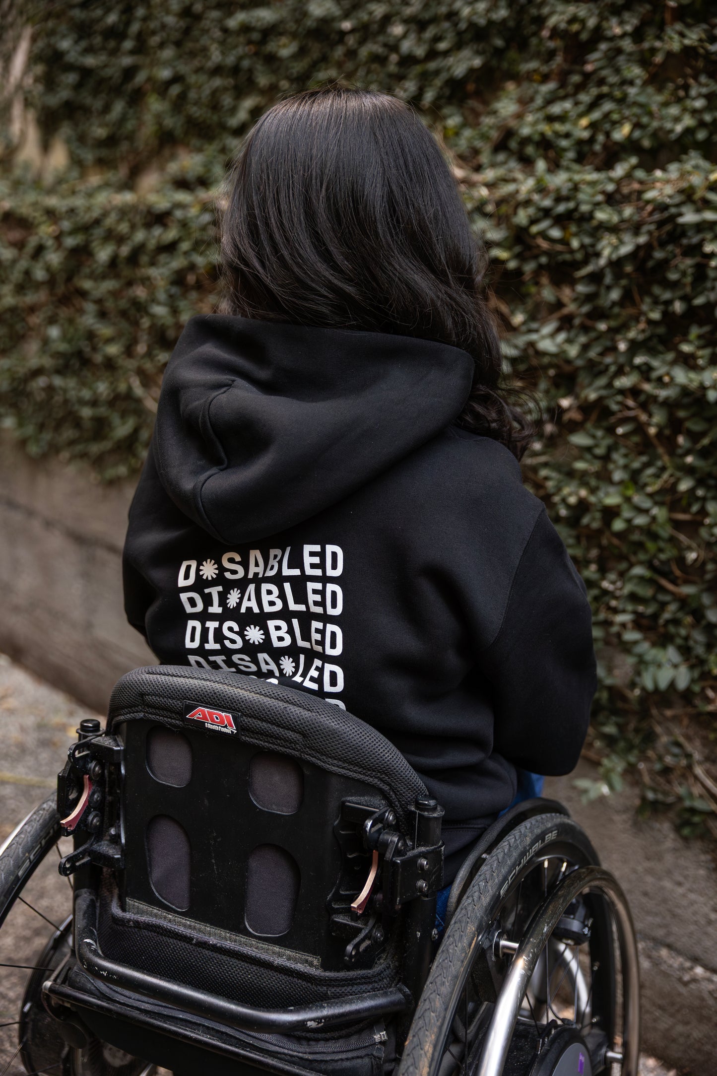 Olivia, sitting in a wheelchair, faces away, wearing a black hoodie with "DISABLED" printed repeatedly in white. Tohuwhetū replace different letters from left to right on each line. Greenery lines the stone path beside them.