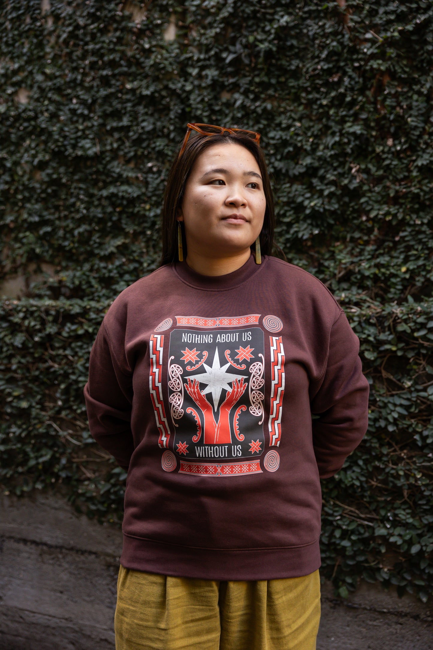 Eda stands against a leafy wall, wearing a dark sweatshirt with the phrase "Nothing About Us Without Us." The graphic features red hands, stars, koru and spirals.