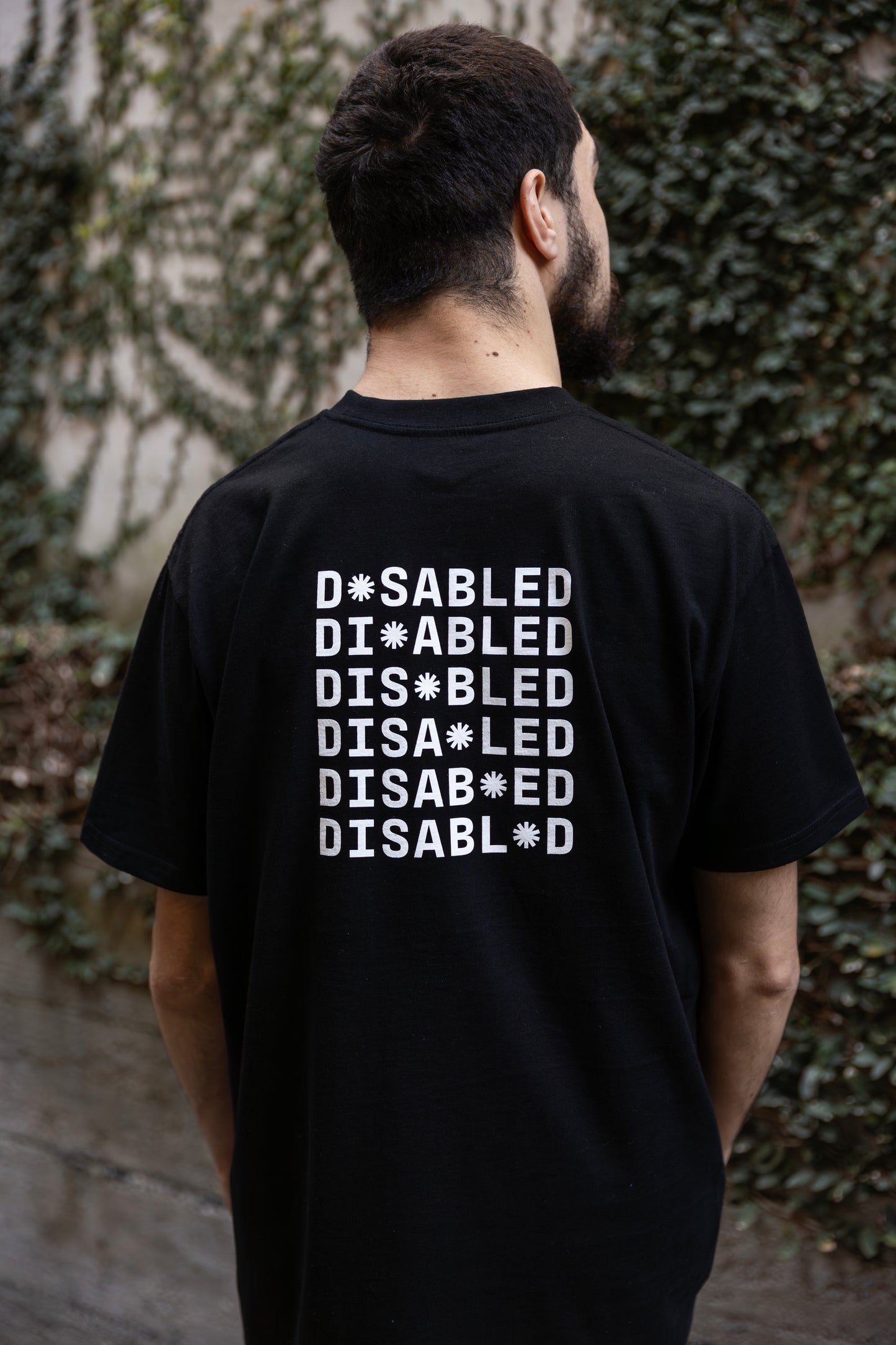 Will stands facing away from the camera, wearing a black T-shirt with "DISABLED" printed multiple times. The white print on the back reads "DISABLED" repeated six times with a tohuwhetū replacing different letters from left to right on each line. He is facing a vine-covered wall.
