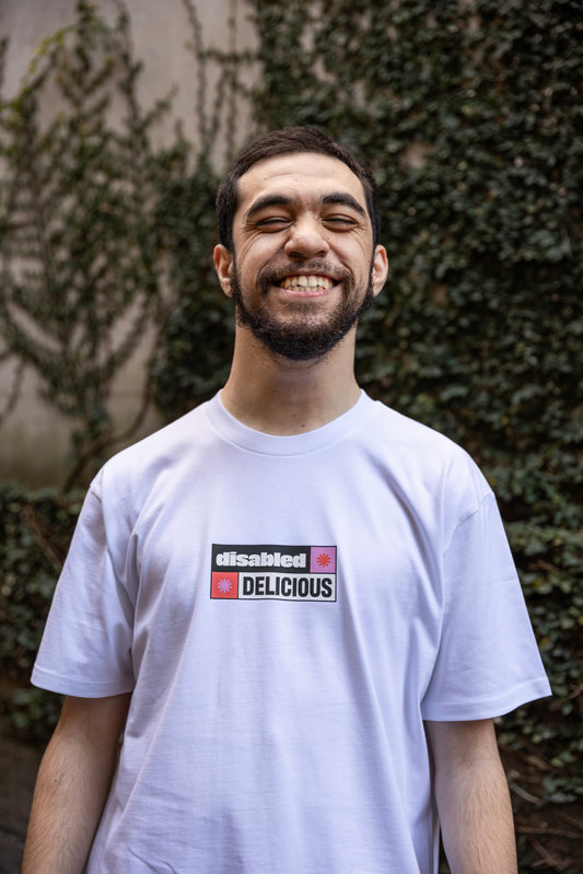 Will wearing a white T-shirt that reads "disabled DELICIOUS." The design features two tohuwhetū and is in pink, red, black and white.