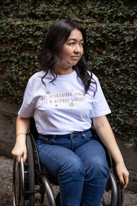 Olivia sits in her wheelchair infront of a leafy wall. She is wearing jeans and a white t-shirt with colourful text which reads "An accessible world is a better world". The design has small designs around it including flowers, a rainbow, a bee, a house and a star.