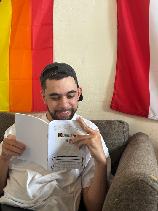 Man in a white shirt and backward cap sits on a brown couch, smiling while reading 'Disabled AF' zine.