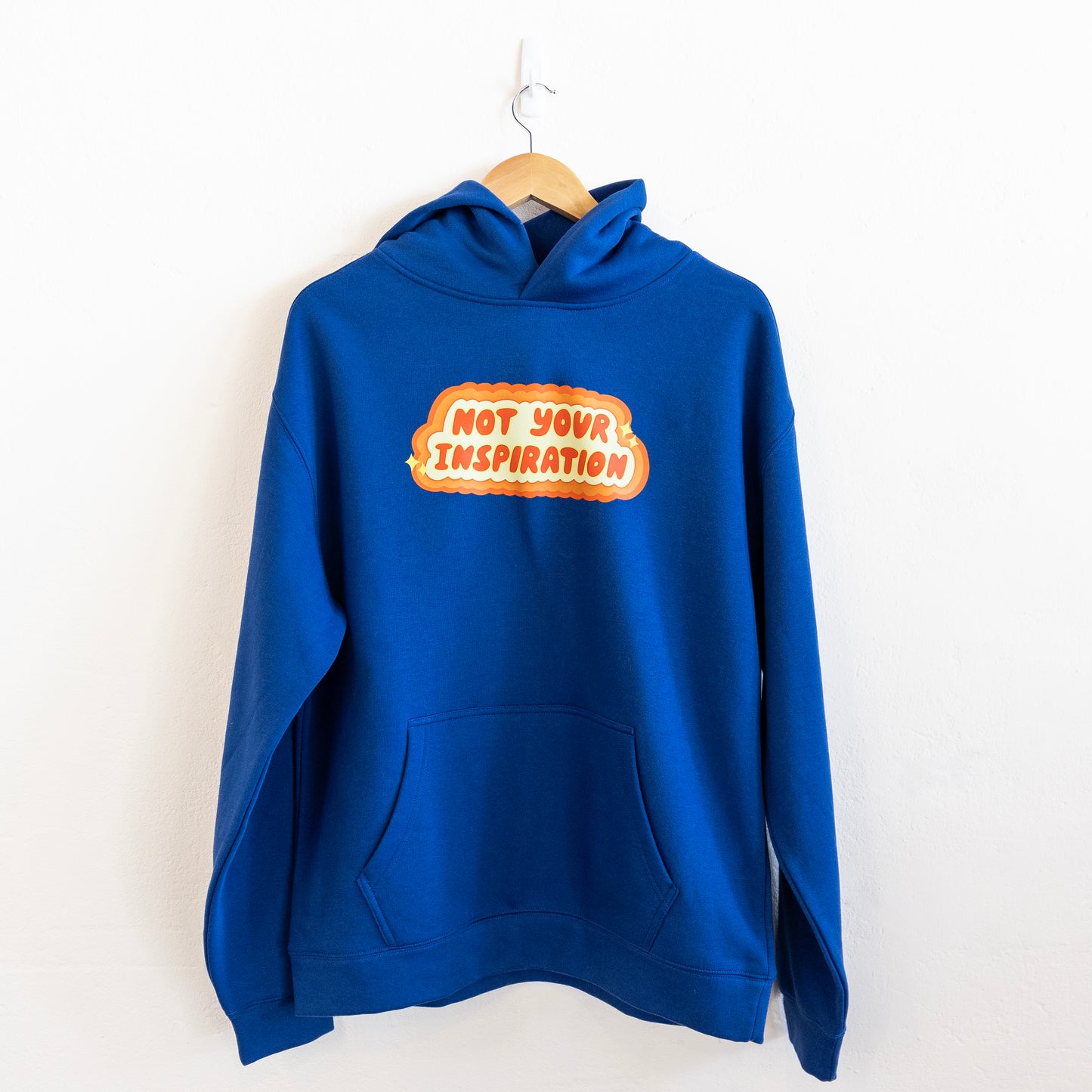 A blue hoodie with a bold, orange and yellow text design on the front reading "Not Your Inspiration" hung up against a white wall.