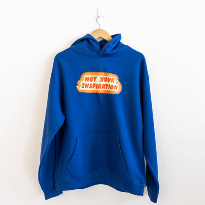 A blue hoodie with a bold, orange and yellow text design on the front reading "Not Your Inspiration" hung up against a white wall.