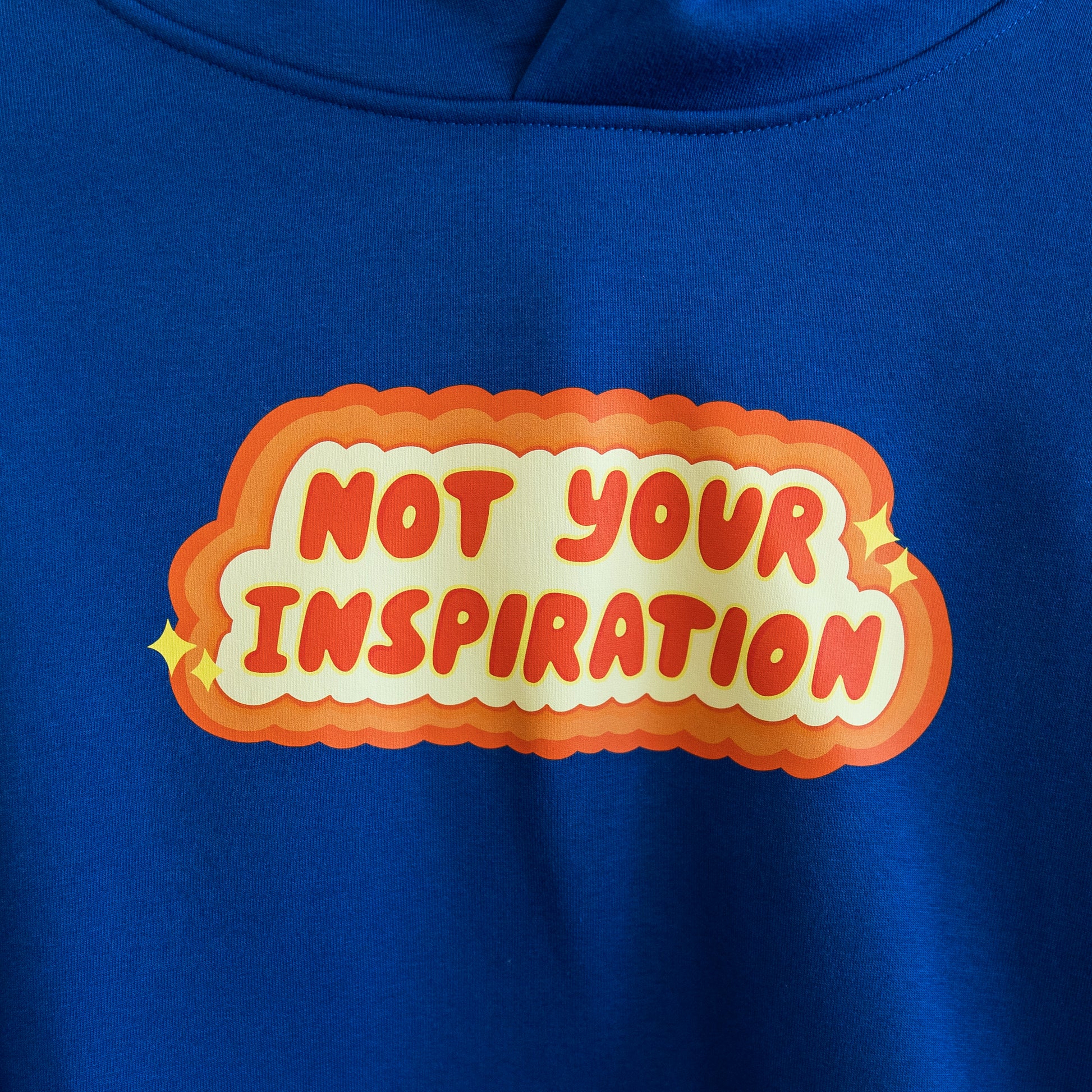 Close up of a blue hoodie with a bold, orange and yellow text design on the front reading "Not Your Inspiration."
