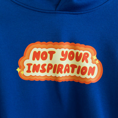 Close up of a blue hoodie with a bold, orange and yellow text design on the front reading "Not Your Inspiration."