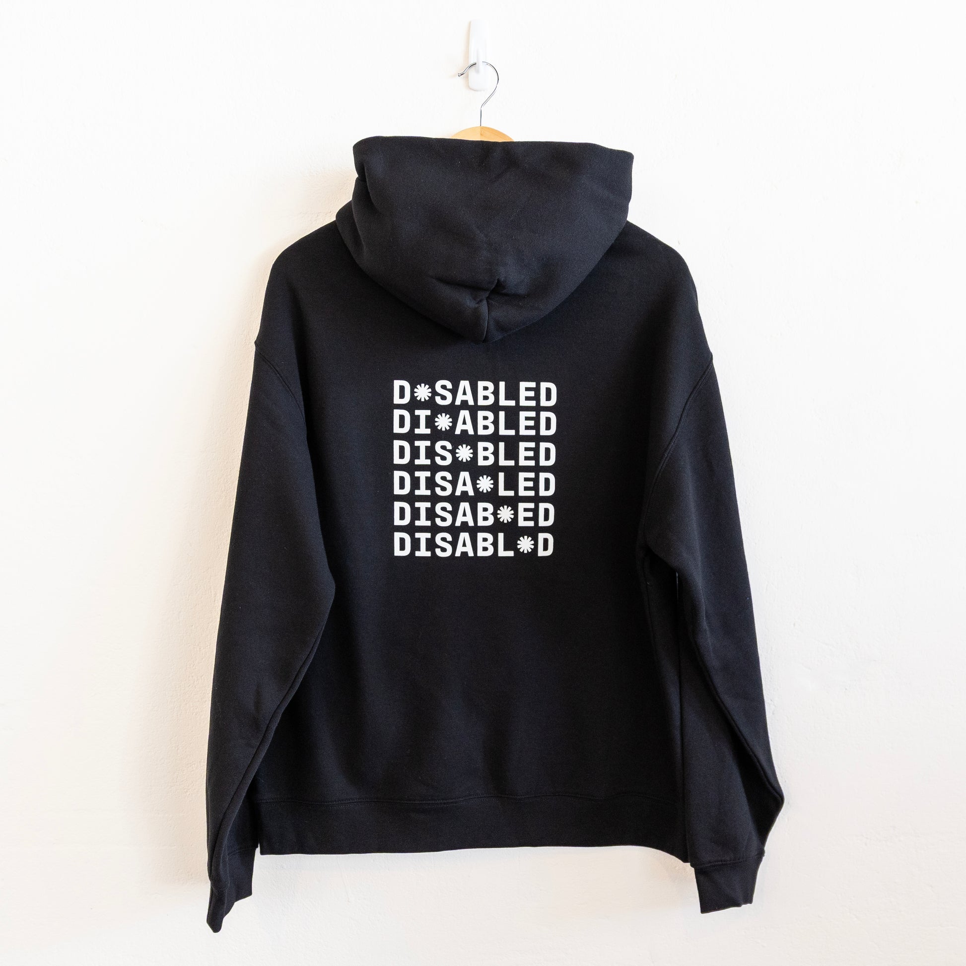 A black hoodie hangs on a white wall, featuring the word "DISABLED" repeated six times in white with a tohuwhetū replacing different letters from left to right on each line.