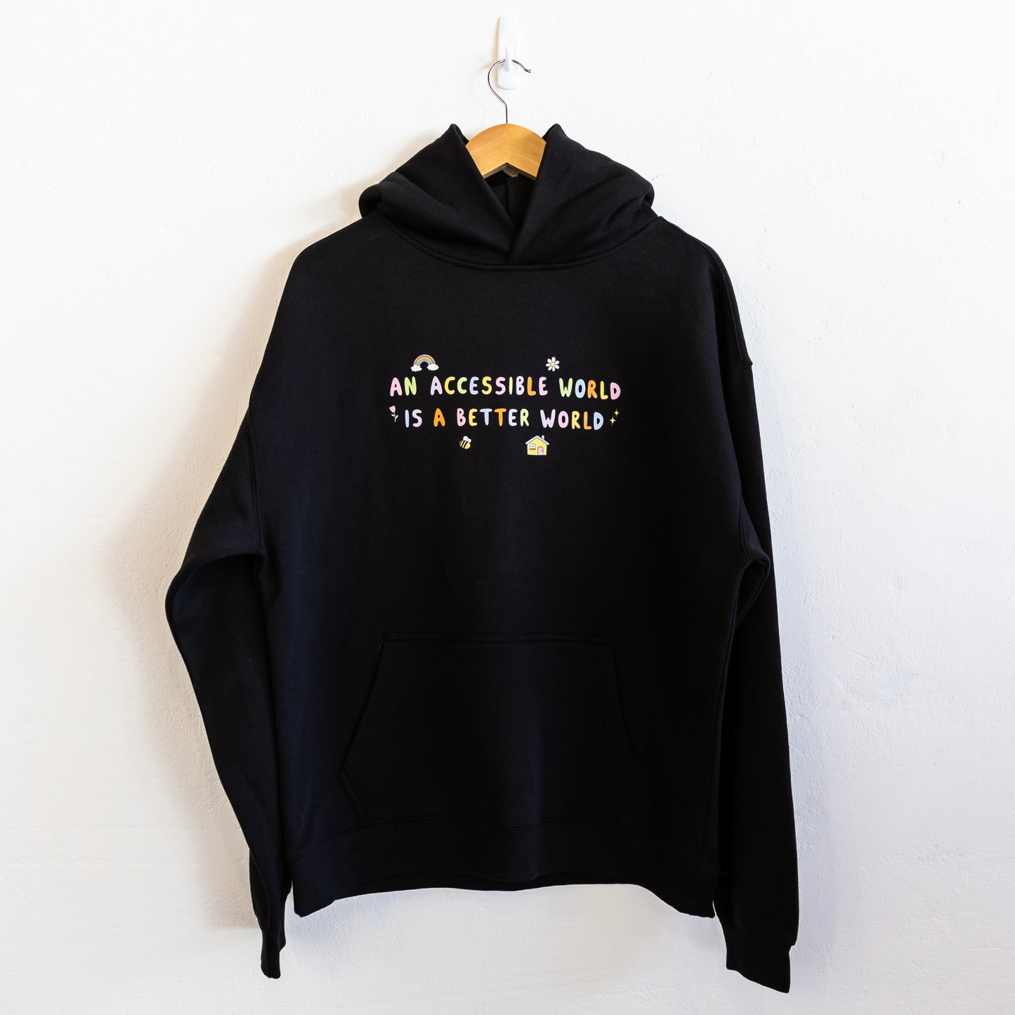 A black hoodie with colourful text which reads "An accessible world is a better world" is hung up against a white wall. The design has small designs around it including flowers, a rainbow, a bee, a house and a star.