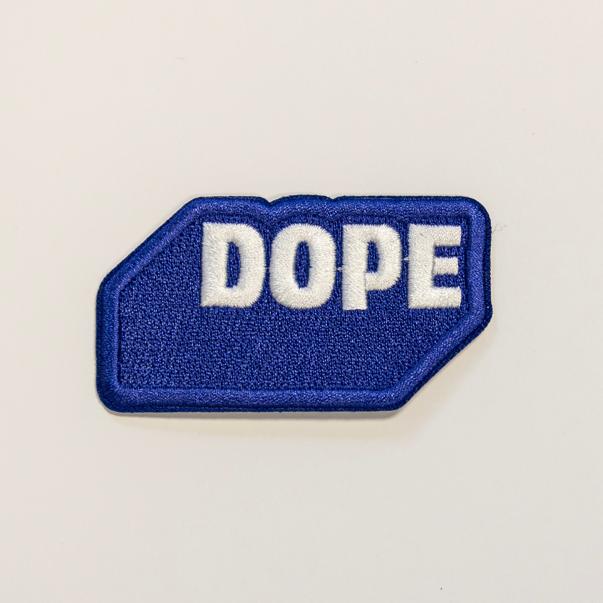 Blue embroidered patch with the word "DOPE" in bold white letters. The patch is shaped as if to mimic a three-dimensional rectangle.