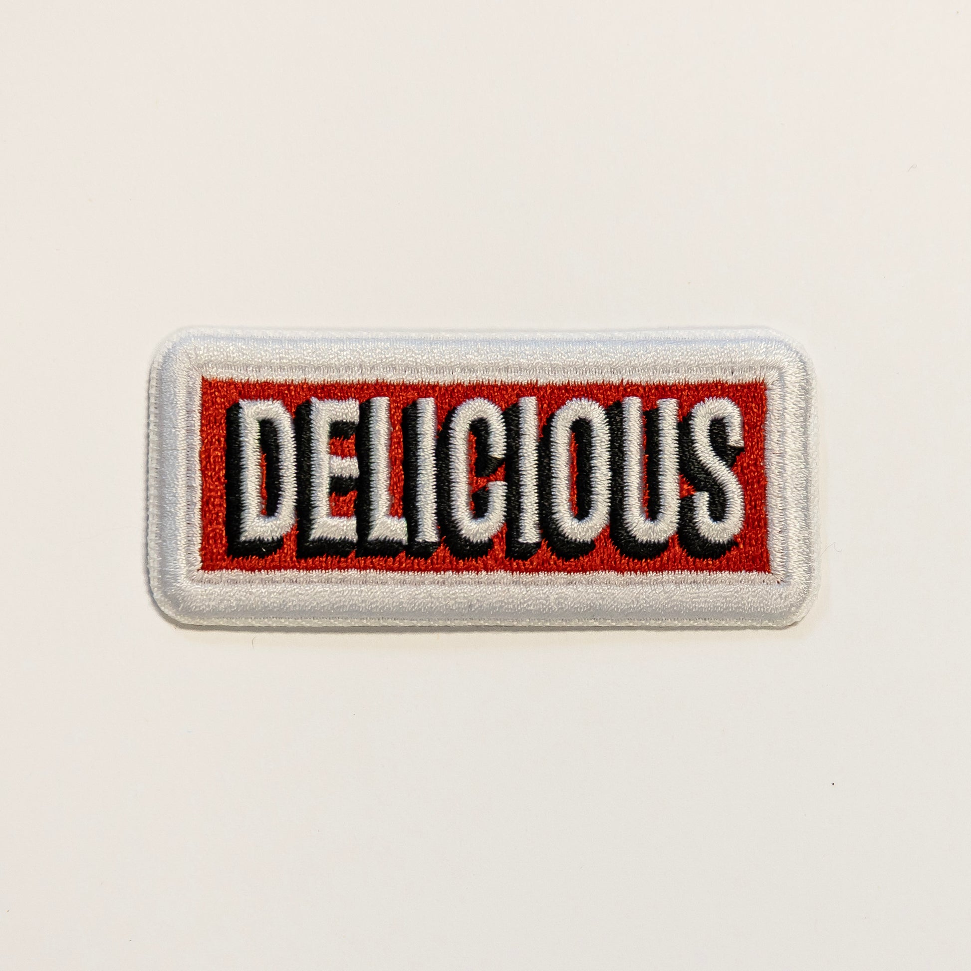 Rectangular embroidered patch with the word "DELICIOUS" in bold white and black letters on a red background, framed by a white border.