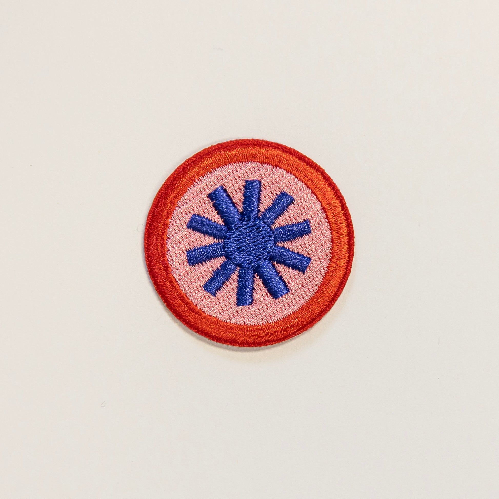 Circular embroidered patch on a light background, featuring a blue tohuwhetū symbol at the center, surrounded by a pink field and red border.