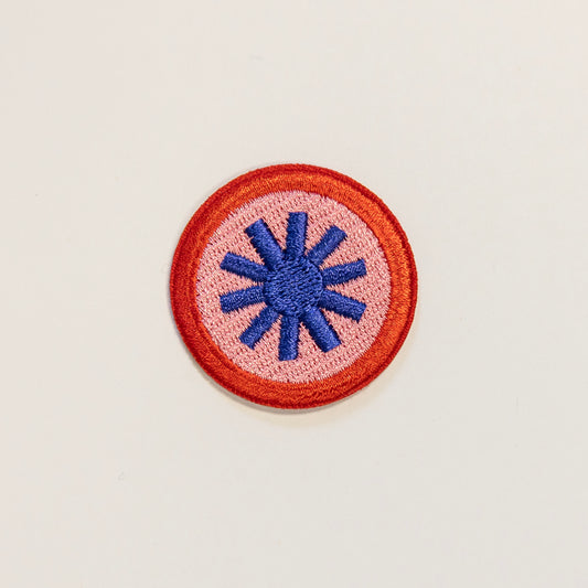 Circular embroidered patch on a light background, featuring a blue tohuwhetū symbol at the center, surrounded by a pink field and red border.