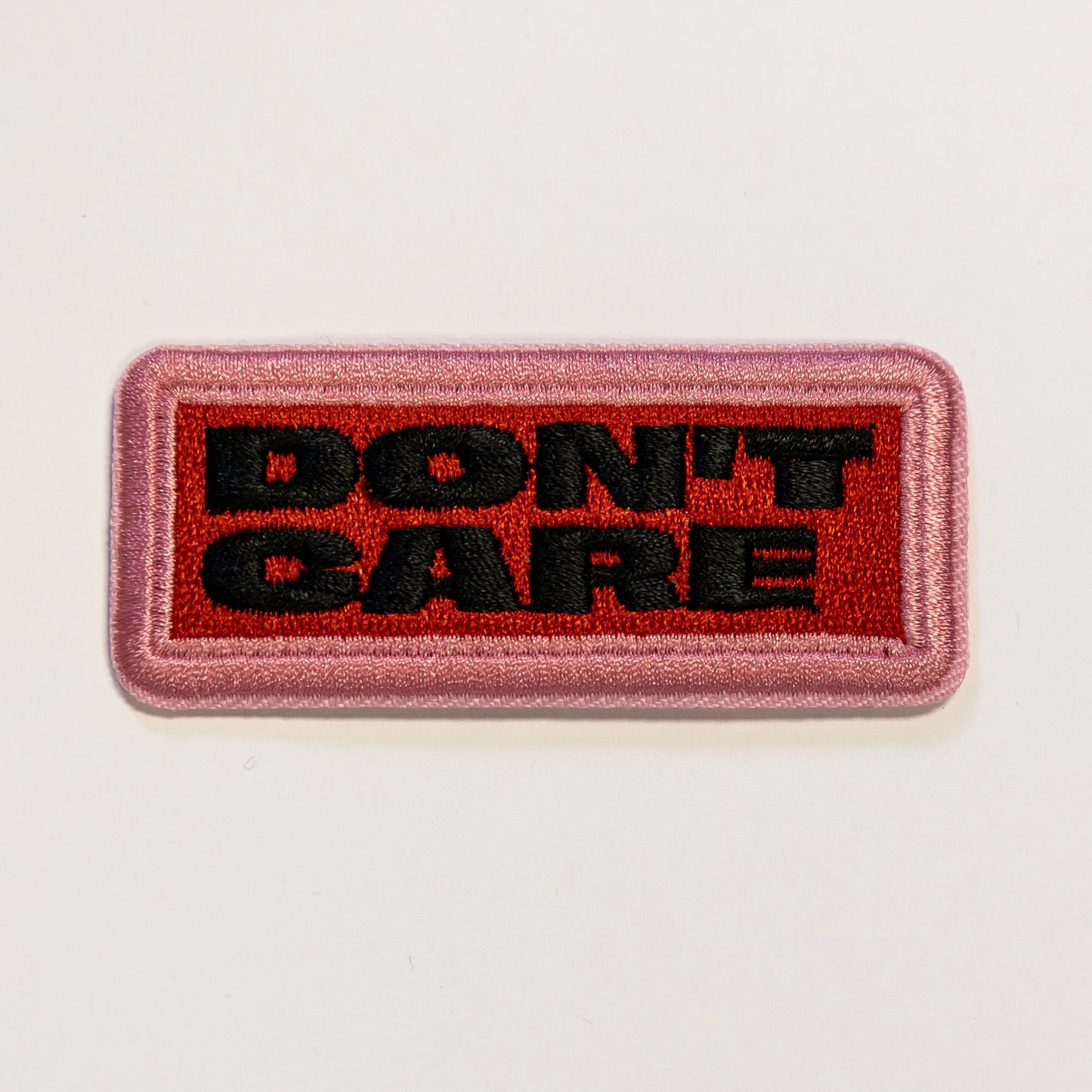 Rectangular embroidered patch with "DON'T CARE" in bold black letters on a red background. The patch has a pink border.