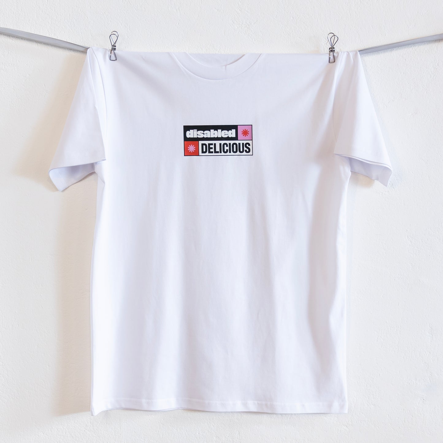 A white T-shirt that reads "disabled DELICIOUS" is hung up on a silver ribbon against a white wall. The design features two tohuwhetū and is in pink, red, black and white.