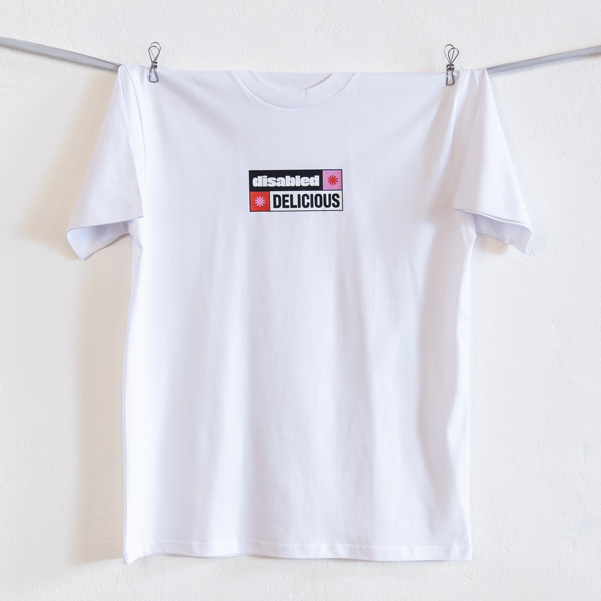 A white T-shirt that reads "disabled DELICIOUS" is hung up on a silver ribbon against a white wall. The design features two tohuwhetū and is in pink, red, black and white.