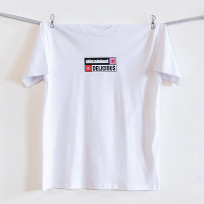 A white T-shirt that reads "disabled DELICIOUS" is hung up on a silver ribbon against a white wall. The design features two tohuwhetū and is in pink, red, black and white.