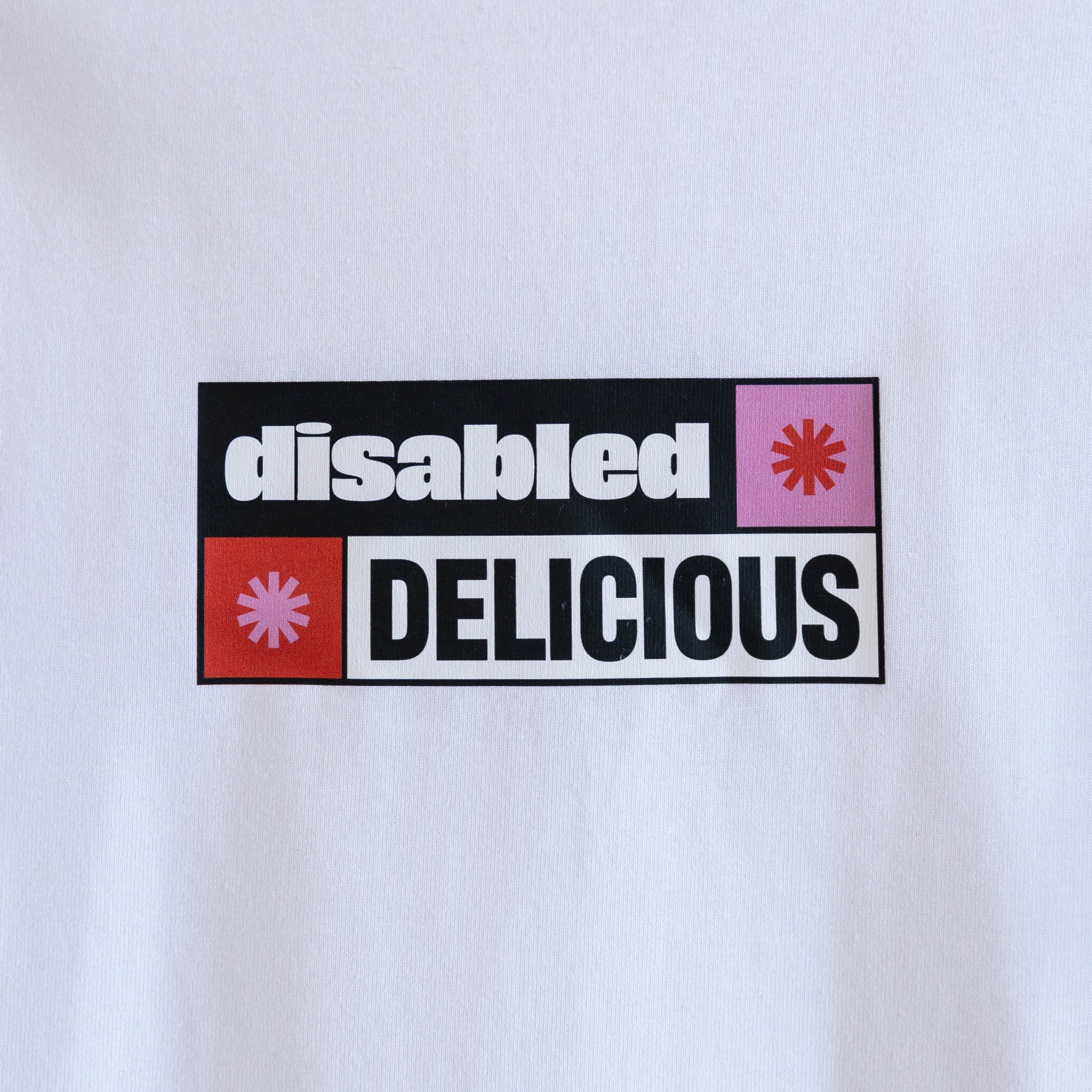 Close up of the print on a white T-shirt that reads "disabled DELICIOUS." The design features two tohuwhetū and is in pink, red, black and white.