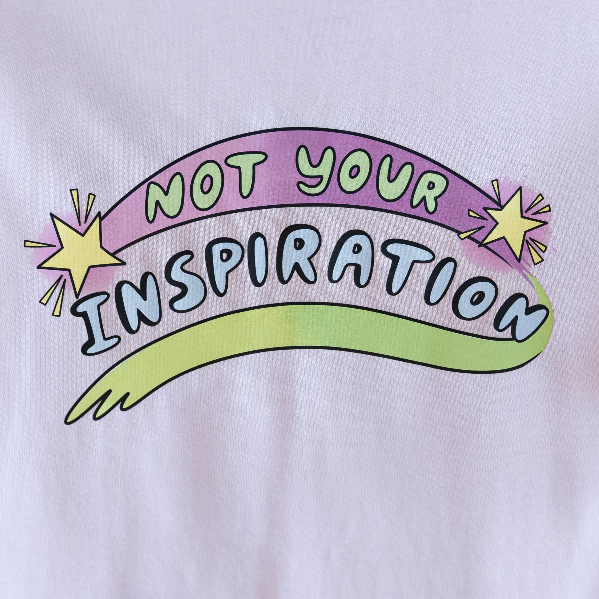 Close up photo of a light pink T-shirt with the phrase "Not Your Inspiration" in colourful letters. The words 'Not your' appear in lime green on a purple swoosh, positioned between two yellow stars. Beneath these words, 'inspiration' appears in all caps, while a second stroke of colour, which appears like a green-yellow paint stroke, provides a boundary of support at the base of the print. 