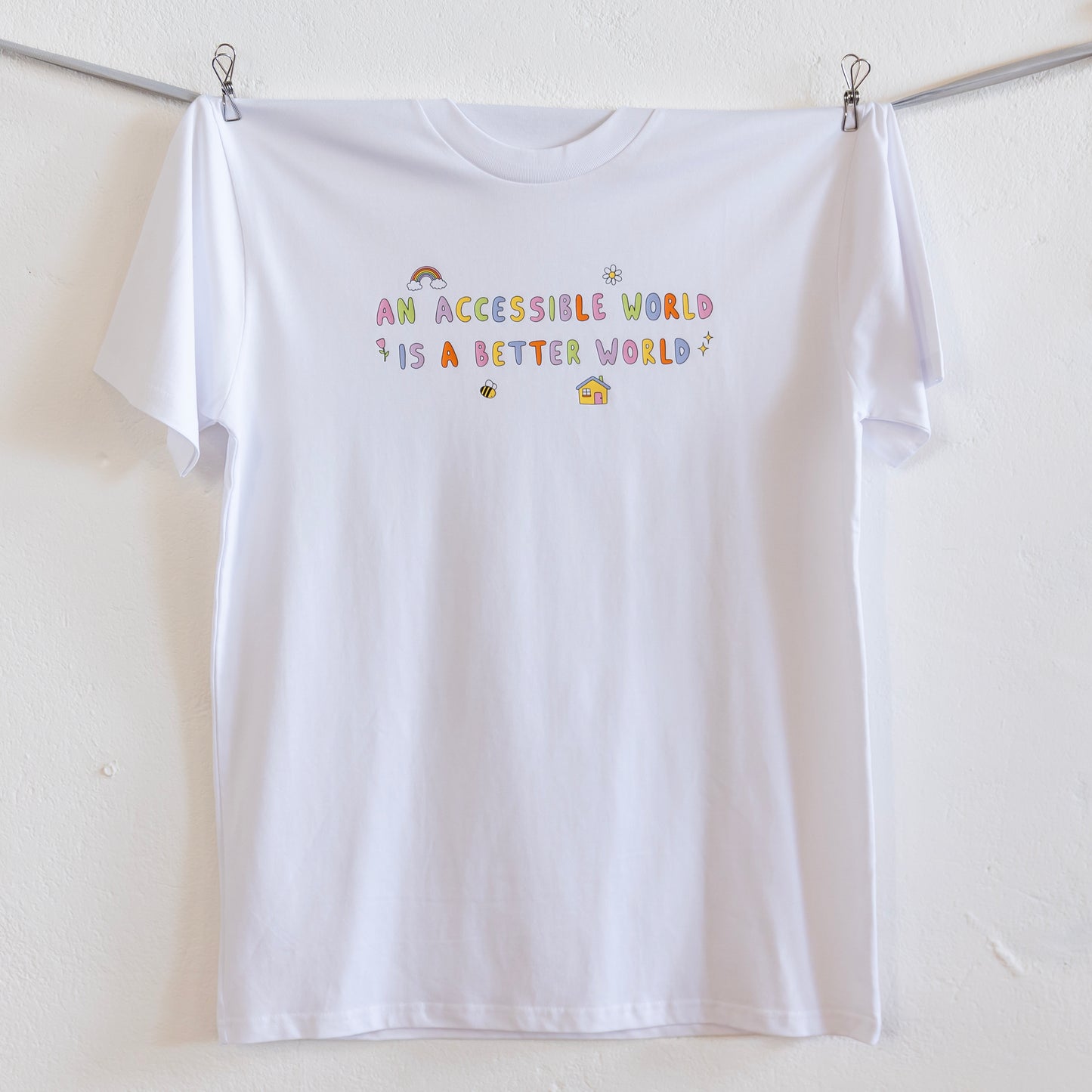 A white t-shirt with colourful text which reads "An accessible world is a better world" is hung up on a grey ribbon against a white wall. The design has small designs around it including flowers, a rainbow, a bee, a house and a star.
