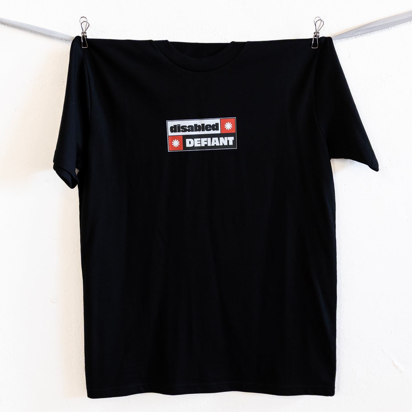 A black T-shirt with "disabled, DEFIANT" in bold text is hung up on a silver ribbon against a white wall. The design is black, red and white, and features two tohuwhetū.