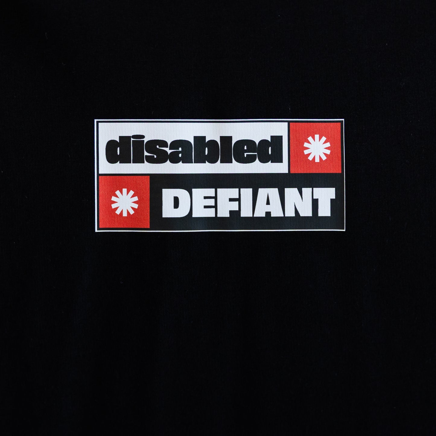 Close up of the print on a black T-shirt with "disabled, DEFIANT" in bold text. The design is black, red and white, and features two tohuwhetū.