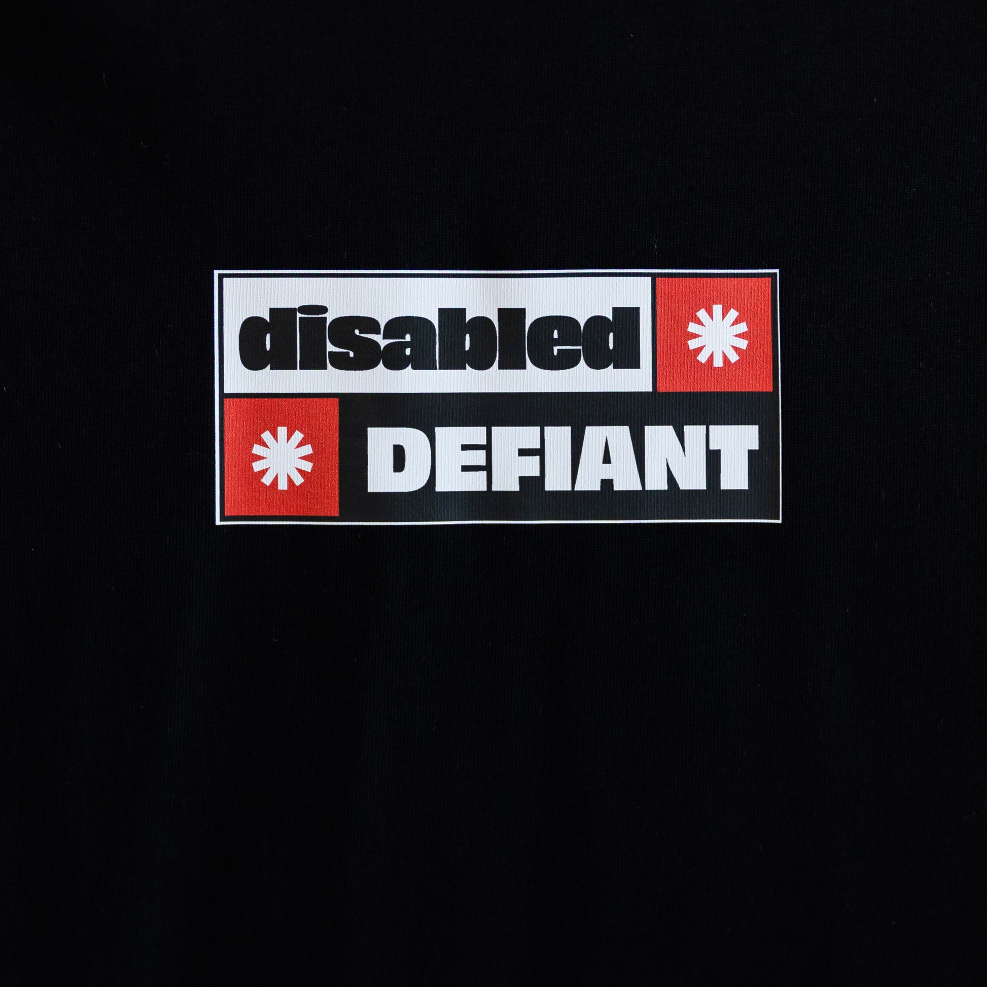 Close up of the print on a black T-shirt with "disabled, DEFIANT" in bold text. The design is black, red and white, and features two tohuwhetū.