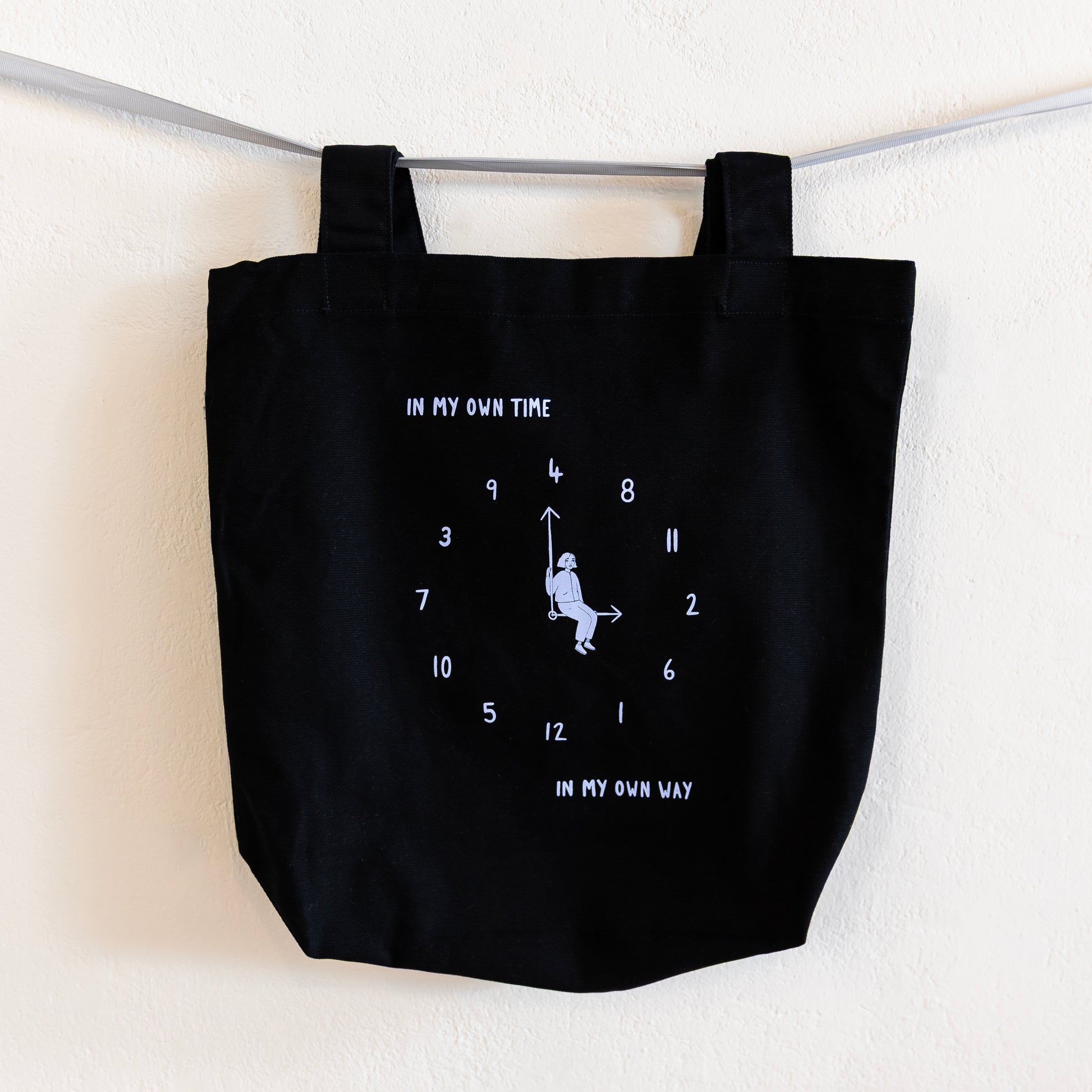 A black tote bag with a light blue print is hung on a silver ribbon against a white wall. The design features an analogue clock, with jumbled numbers and a person sitting on the clock hands. Above the design reads "IN MY OWN TIME" and below reads "IN MY OWN WAY".