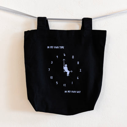 A black tote bag with a light blue print is hung on a silver ribbon against a white wall. The design features an analogue clock, with jumbled numbers and a person sitting on the clock hands. Above the design reads "IN MY OWN TIME" and below reads "IN MY OWN WAY".