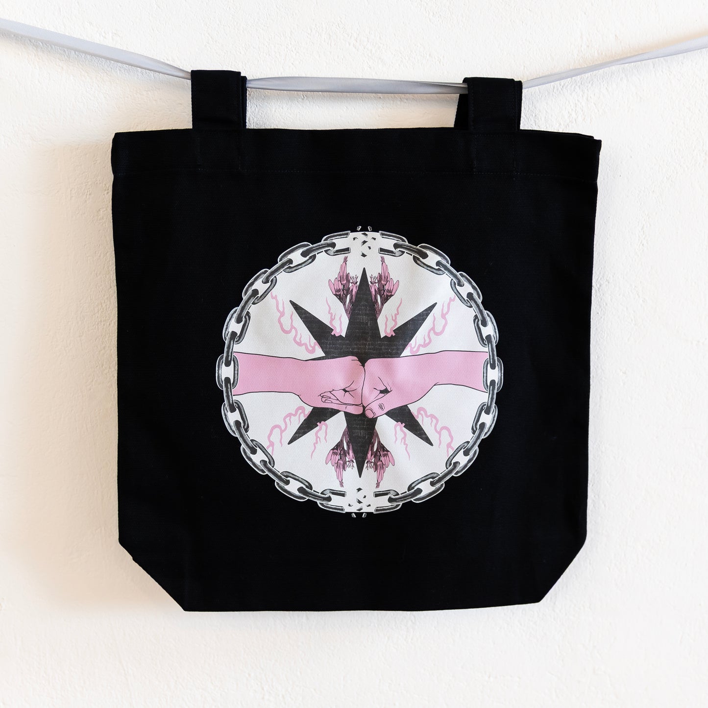 A black tote bag is hung up on a silver ribbon against a white wall. The bag features a central circular design of two pink fists bumping inside a chain circle with breaks at the top and bottom. The inside of the circle has a beige background, and the fists are in-front of a black star with pink flames and flowers branching off of it.