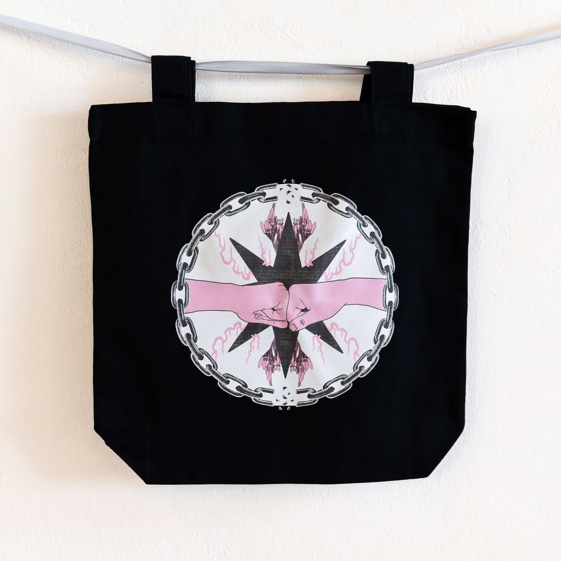 A black tote bag is hung up on a silver ribbon against a white wall. The bag features a central circular design of two pink fists bumping inside a chain circle with breaks at the top and bottom. The inside of the circle has a beige background, and the fists are in-front of a black star with pink flames and flowers branching off of it.
