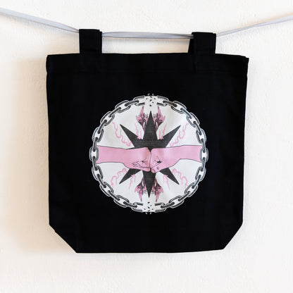 A black tote bag is hung up on a silver ribbon against a white wall. The bag features a central circular design of two pink fists bumping inside a chain circle with breaks at the top and bottom. The inside of the circle has a beige background, and the fists are in-front of a black star with pink flames and flowers branching off of it.