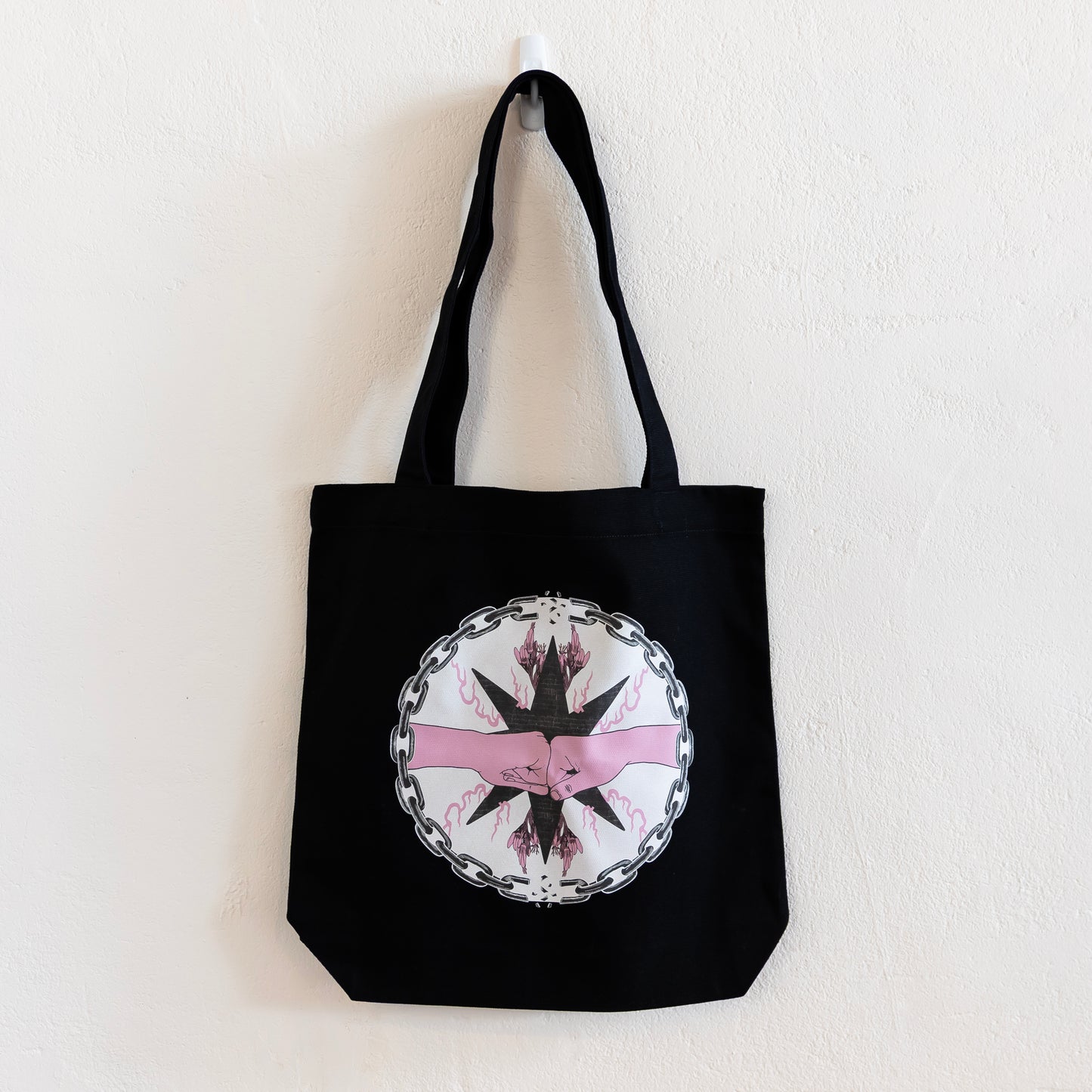 A black tote bag is hung up on a white wall. The bag features a central circular design of two pink fists bumping inside a chain circle with breaks at the top and bottom. The inside of the circle has a beige background, and the fists are in-front of a black star with pink flames and flowers branching off of it.