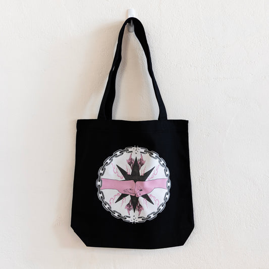 A black tote bag is hung up on a white wall. The bag features a central circular design of two pink fists bumping inside a chain circle with breaks at the top and bottom. The inside of the circle has a beige background, and the fists are in-front of a black star with pink flames and flowers branching off of it.