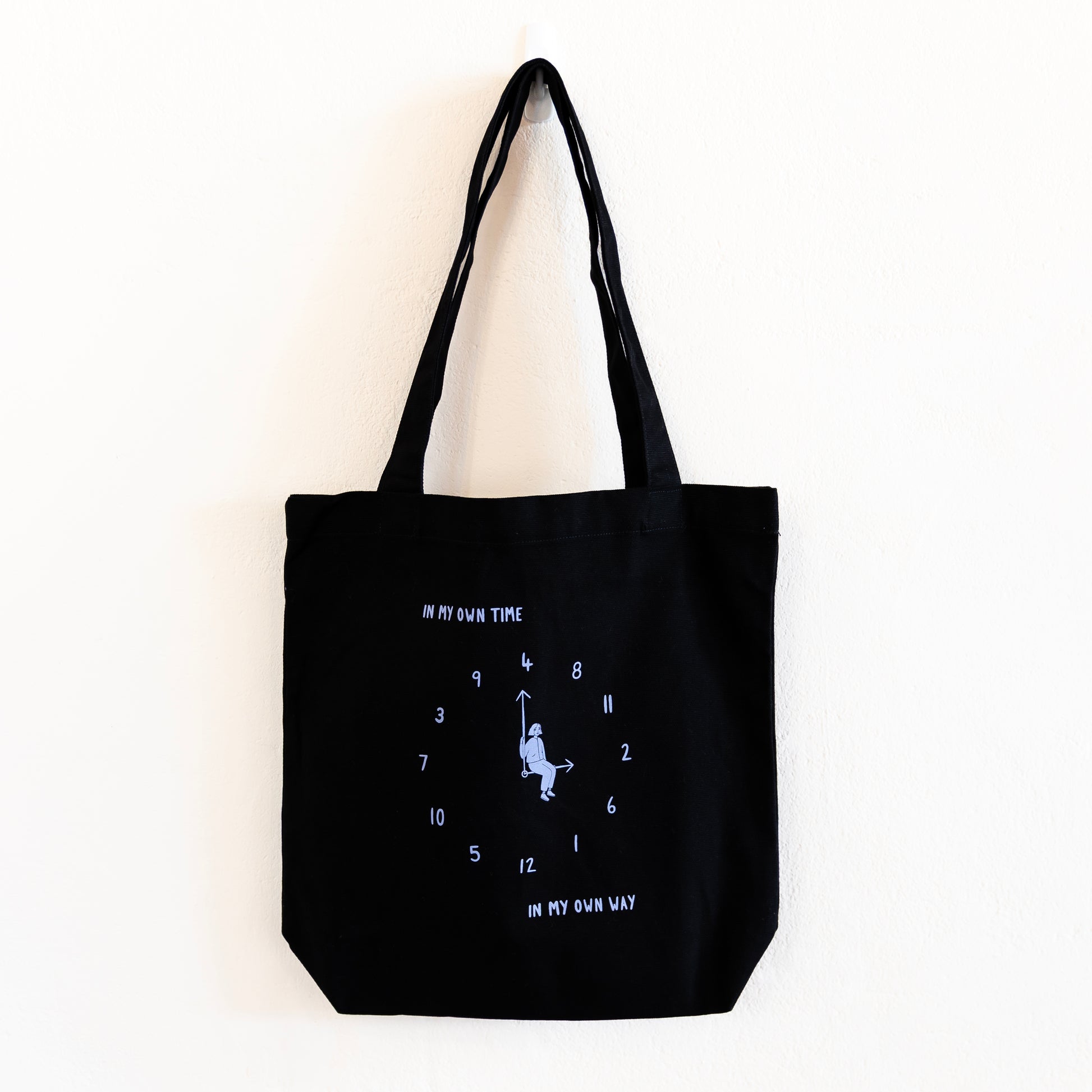 A black tote bag with a light blue print is hung against a white wall. The design features an analogue clock, with jumbled numbers and a person sitting on the clock hands. Above the design reads "IN MY OWN TIME" and below reads "IN MY OWN WAY".