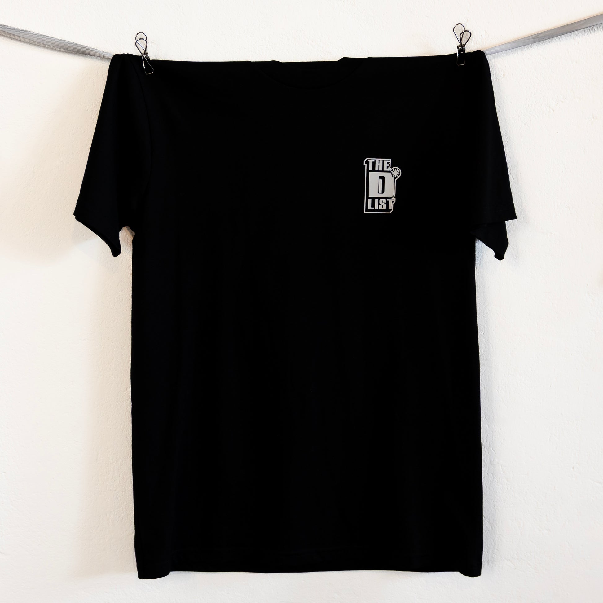 Black t-shirt hung up on a silver ribbon against a white background. Features white "The D*List" logo on the left chest.