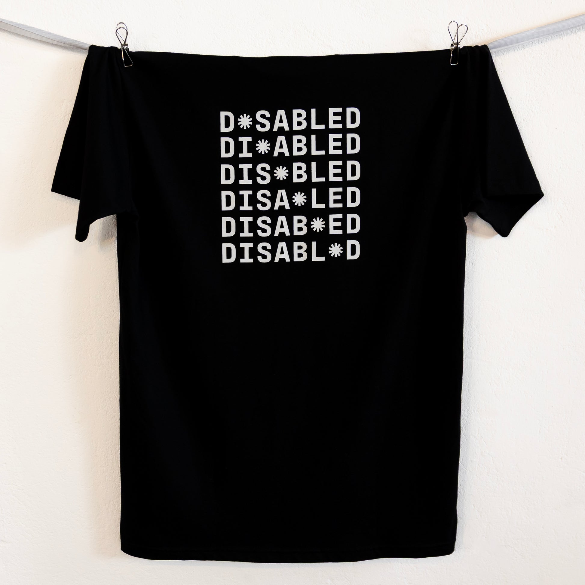 Black t-shirt hung up on a silver ribbon against a white background. The tee features the word "DISABLED" repeated six times in white with a tohuwhetū replacing different letters from left to right on each line.