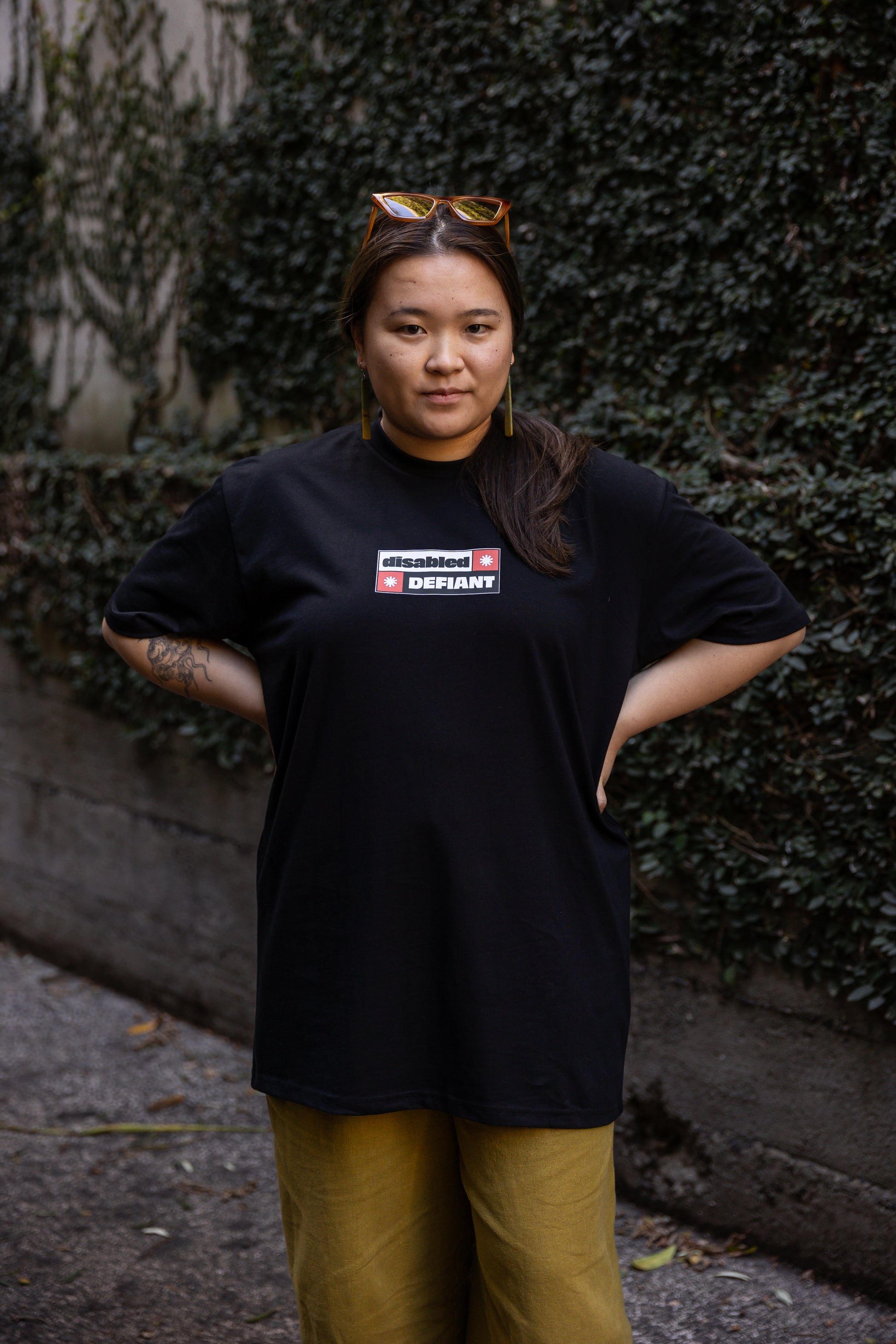 Eda stands in-front of a leafy wall outdoors, wearing a black T-shirt, untucked, with "disabled, DEFIANT" in bold text stands outdoors. The design is black, red and white, and features two tohuwhetū.